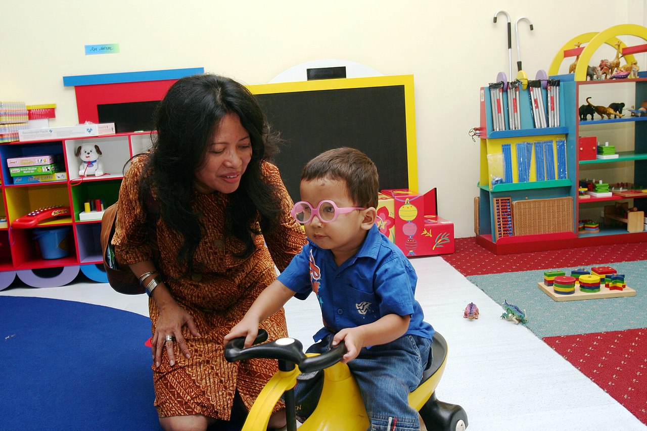 Guru mendampingi seorang anak bermain di ruang kelas berwarna cerah